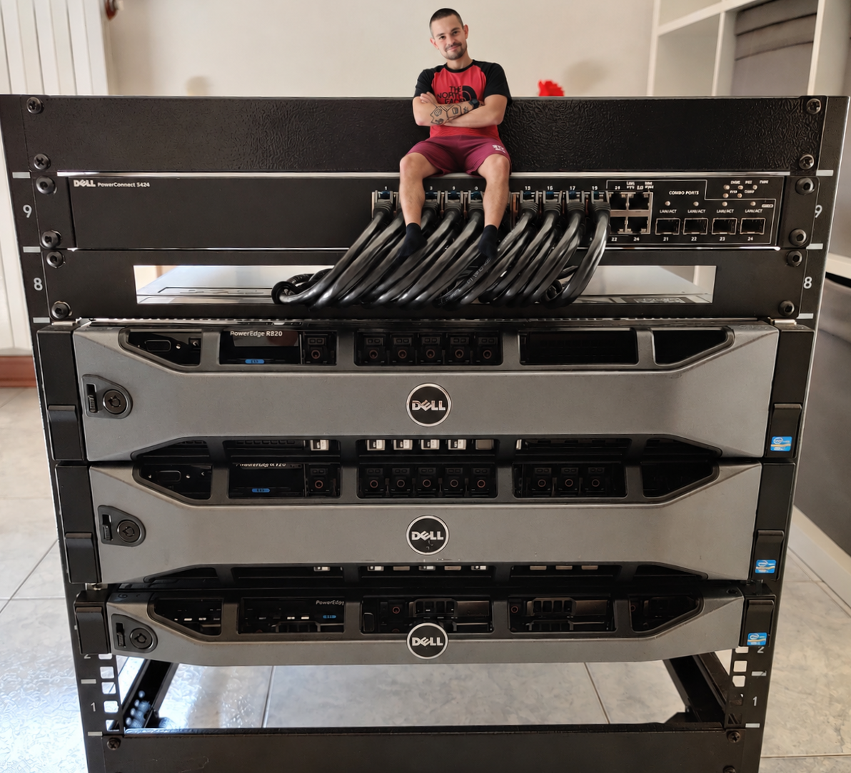 An open-frame server rack in a home lab: a Dell PowerConnect 5424 switch with a bundle of network cables at the top, three Dell PowerEdge R820 servers stacked below it, and a person sitting on top of the rack.